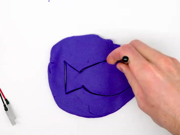 Hand drawing a fish outline on flattened purple dough with a dough tool